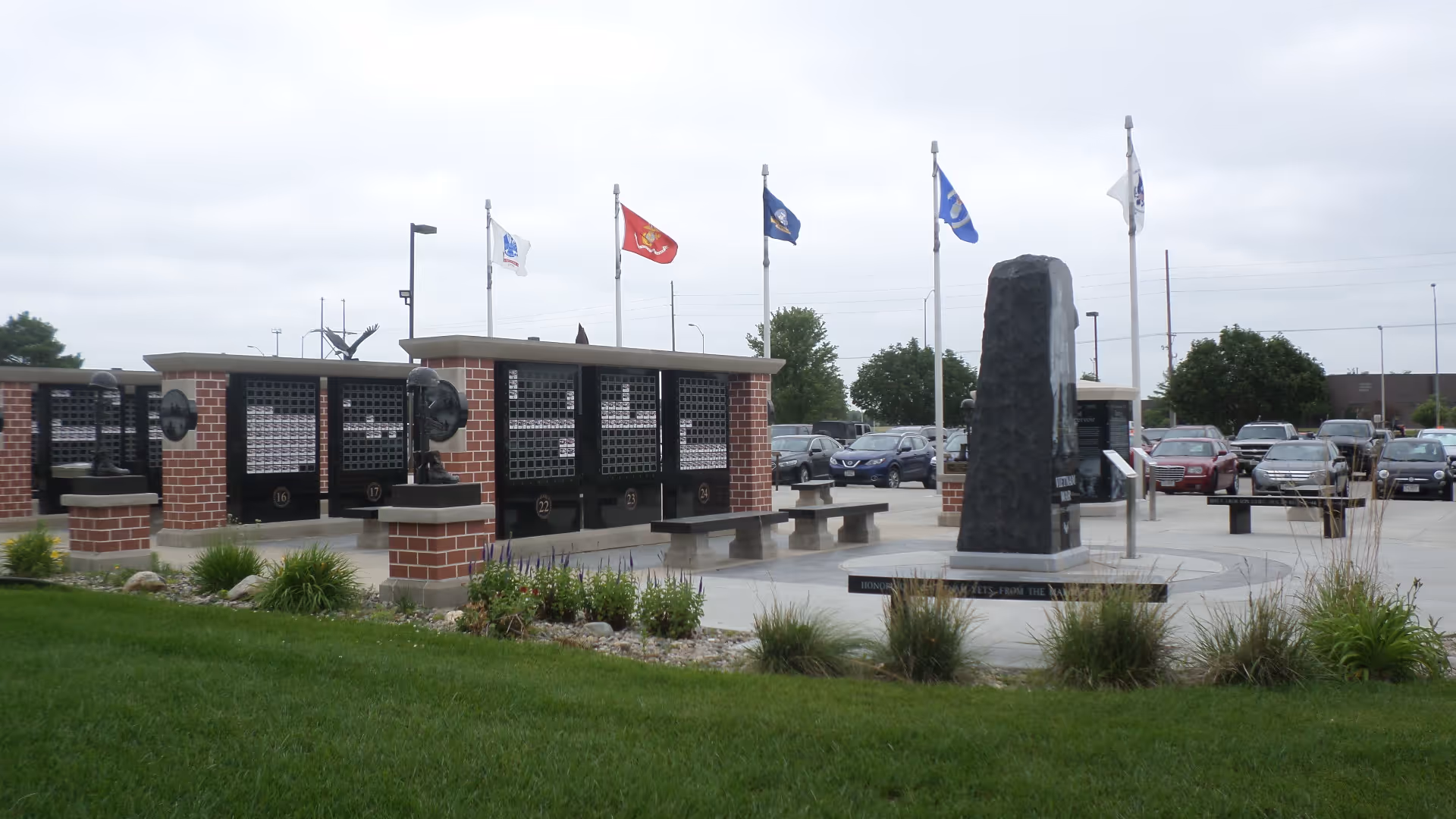 Outdoor memorial area at Norfolk Veterans' Home featuring a tall dark stone monument surrounded by benches and brick structures with plaques. Several flags are flying on flagpoles behind the monument, and there is a parking lot with cars in the background under a cloudy sky.