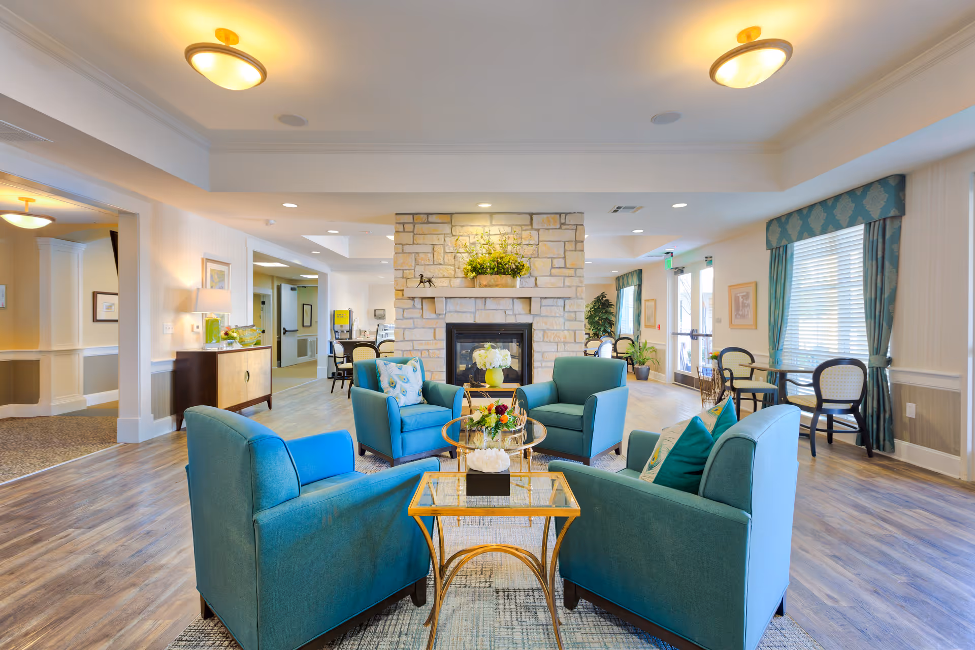 Bright communal lounge with teal armchairs arranged around a glass coffee table facing a stone fireplace.