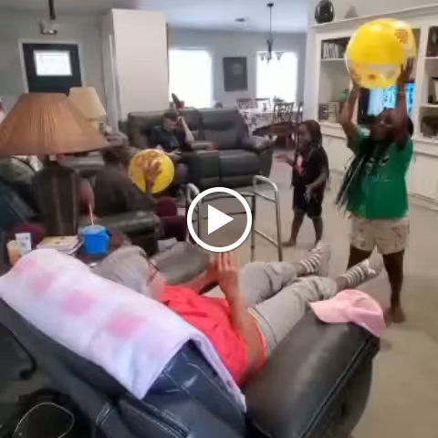 An elderly person reclines on a leather chair with a pink towel over the backrest, playing with a yellow beach ball with two children standing nearby in a living room. The room has a sofa, a lamp, and a dining area visible in the background.