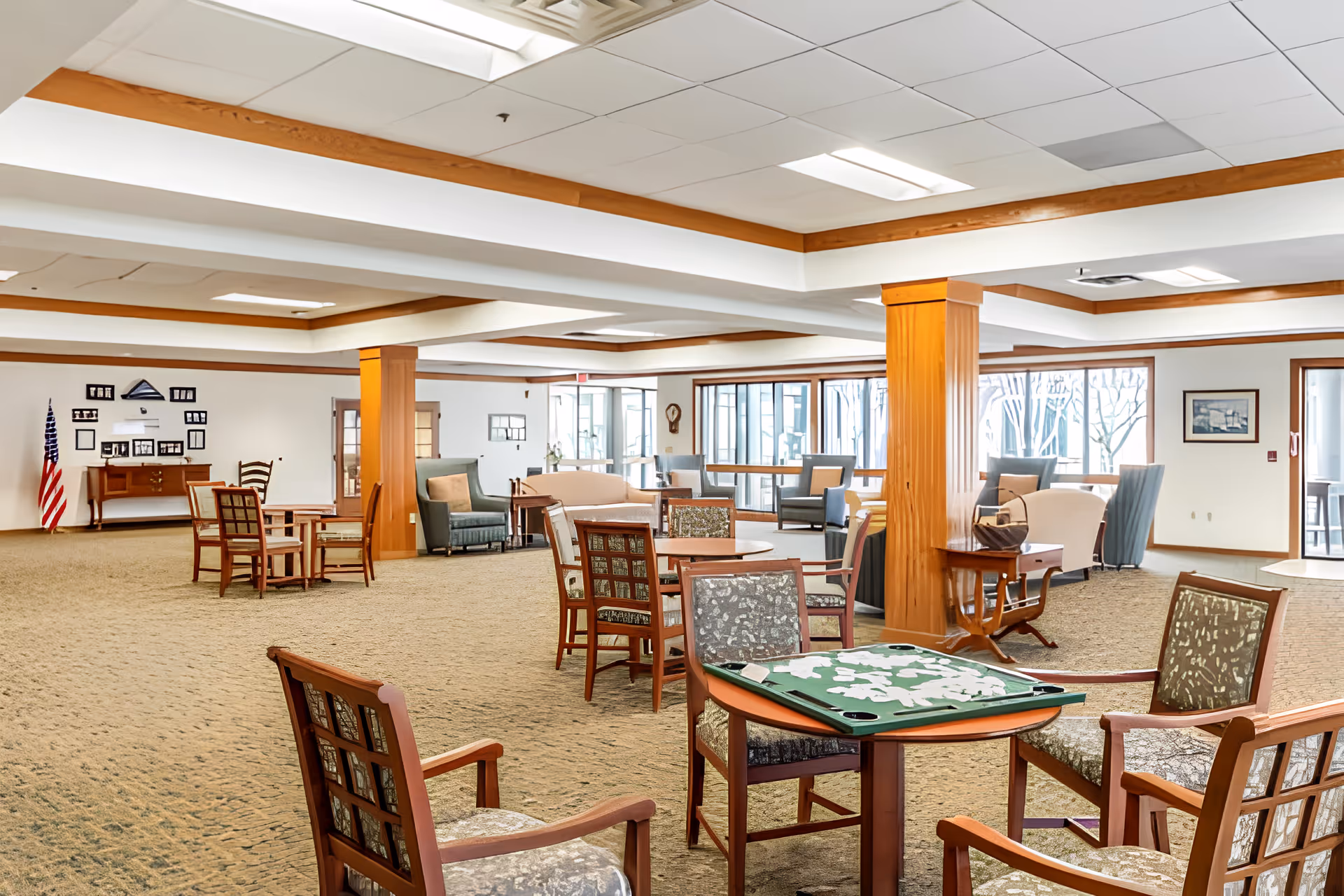 A spacious common area in an assisted living facility with multiple seating arrangements including wooden chairs and tables, armchairs, and sofas. The room features carpeted floors, wooden columns, large windows letting in natural light, and a ceiling with recessed lighting. A game board with pieces is set on one of the tables in the foreground.