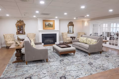 A spacious and well-lit living room with two gray sofas facing each other, a cushioned ottoman in the center with a wooden tray on top, two patterned armchairs near the walls, a fireplace with a framed painting above it, and two ornate mirrors on either side of the fireplace. The room has a large patterned area rug and wooden flooring, with French doors visible on the right side.