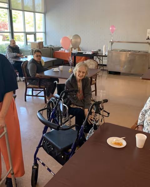 Several elderly individuals seated at tables in a bright room with large windows. One elderly woman is sitting in a wheelchair near the foreground, smiling. There are balloons tied to a table in the background and a serving station along the wall.