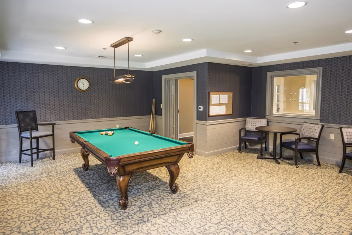 A recreational room with a pool table in the center, a high chair against the wall, a small round table with three chairs, patterned carpet, and dark patterned wallpaper with wainscoting. A clock is mounted on the wall above the pool cues in the corner.