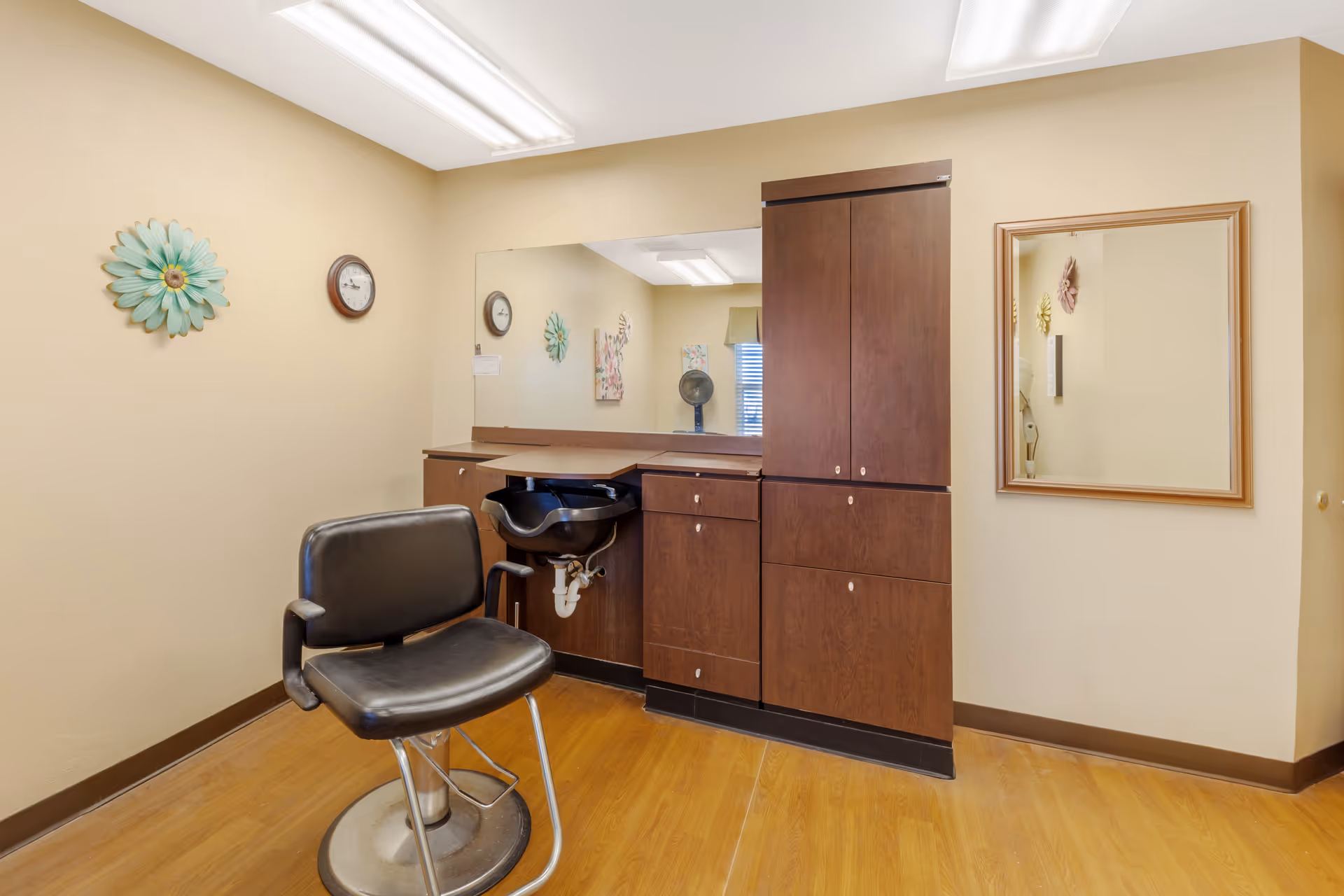 Empty salon styling station with a black barber chair, shampoo sink, large mirror, and dark wood cabinets.