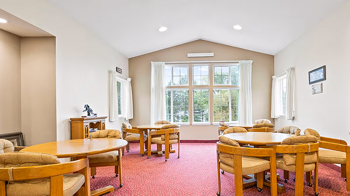 A bright room with large windows letting in natural light, featuring several round wooden tables surrounded by cushioned chairs on a red carpeted floor. The walls are painted beige and white curtains frame the windows.