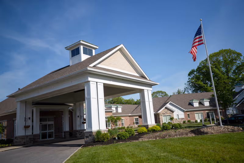 Exterior view of a senior living facility building with a covered entrance, well-maintained landscaping, and an American flag on a flagpole against a blue sky.