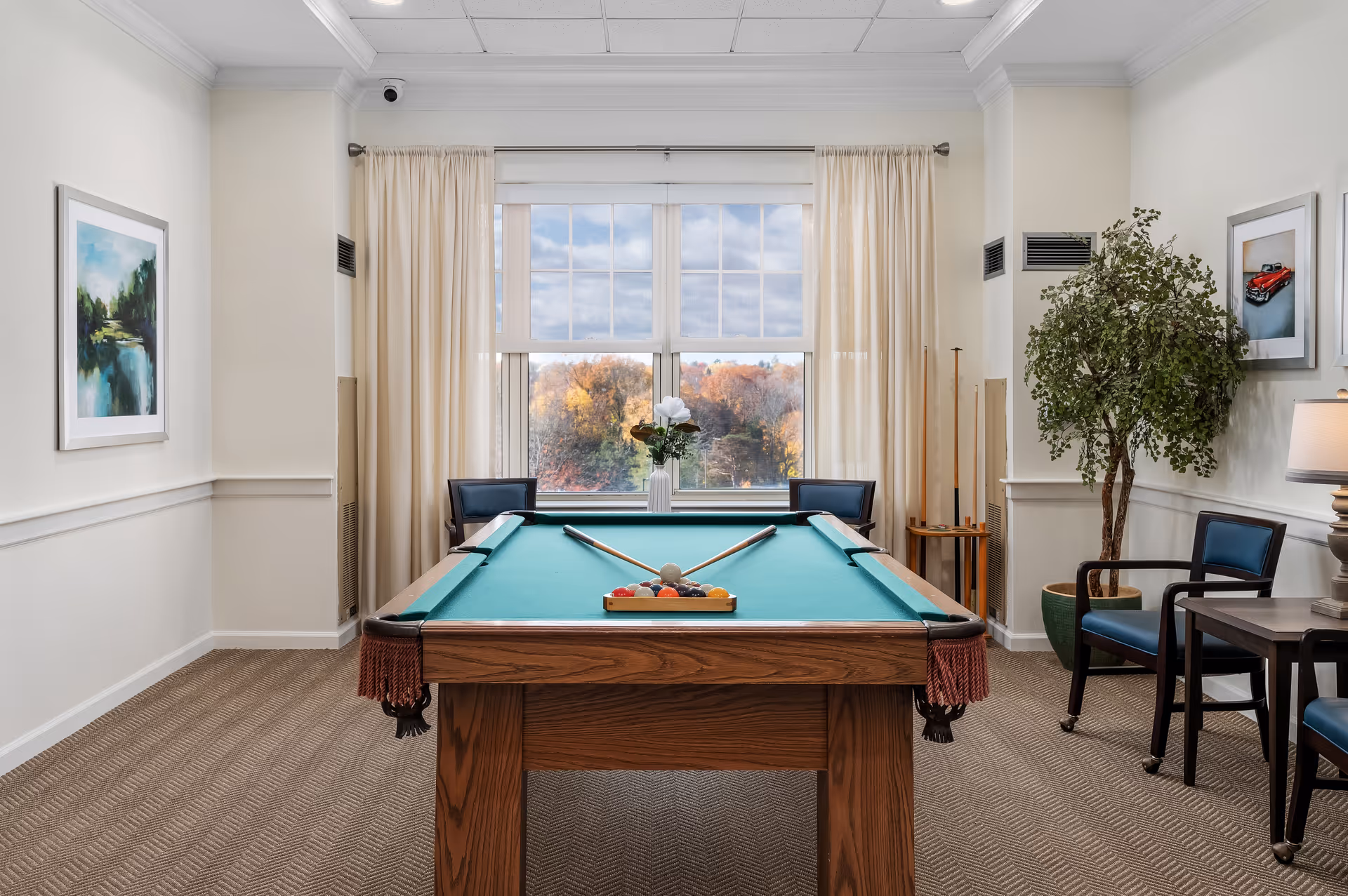 A bright room with a pool table in the center, two pool cues resting on the table, and a rack of billiard balls arranged at one end. The room has beige walls, carpeted floor, and a large window with cream-colored curtains showing an outdoor view with trees. There are two chairs on either side of the window, a potted plant, framed artwork on the walls, and a small table with a lamp on the right side.