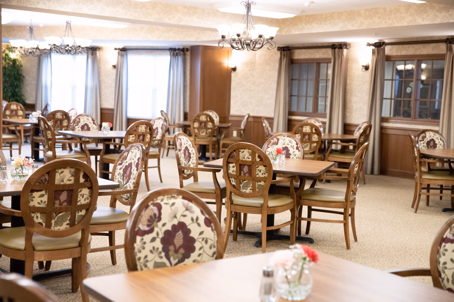A bright and spacious dining room with multiple wooden tables and chairs featuring floral upholstery. The room has large windows with curtains, chandeliers hanging from the ceiling, and small flower arrangements on each table.