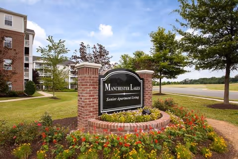 Outdoor view of the entrance sign for Manchester Lakes Senior Apartment Living, surrounded by a landscaped garden with colorful flowers and greenery, with apartment buildings and trees in the background under a partly cloudy sky.