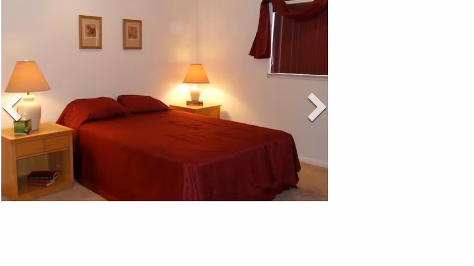 A bedroom with a double bed covered in a dark red bedspread and matching pillows. There are two wooden nightstands on either side of the bed, each with a lamp. The wall behind the bed has two framed pictures, and there is a window with dark red curtains on the right side.