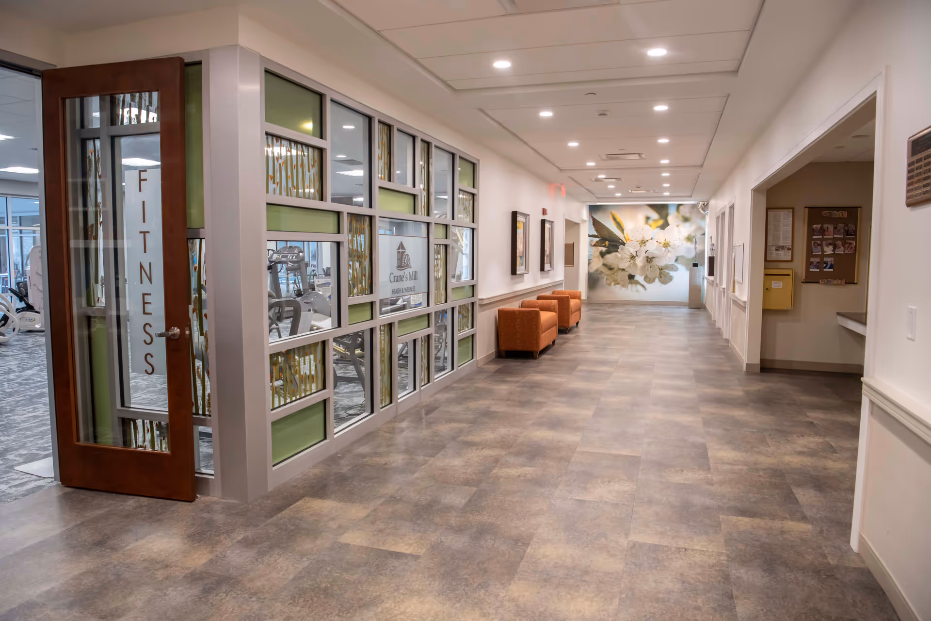 A wide hallway in a senior living facility with a fitness room on the left behind glass walls and a wooden door labeled 'FITNESS'. The hallway has a tiled floor, white walls, recessed ceiling lights, and orange armchairs along the right wall. At the end of the hallway is a large floral mural.