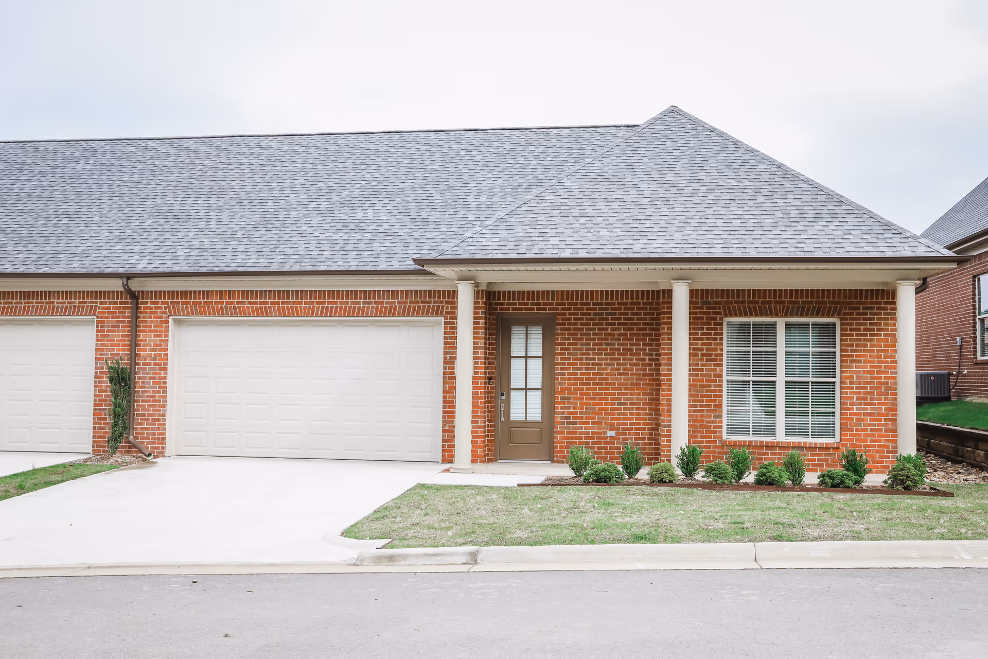 Front exterior of a single-story brick building with a double garage, central entry door, window, and small landscaped lawn under a pitched roof.