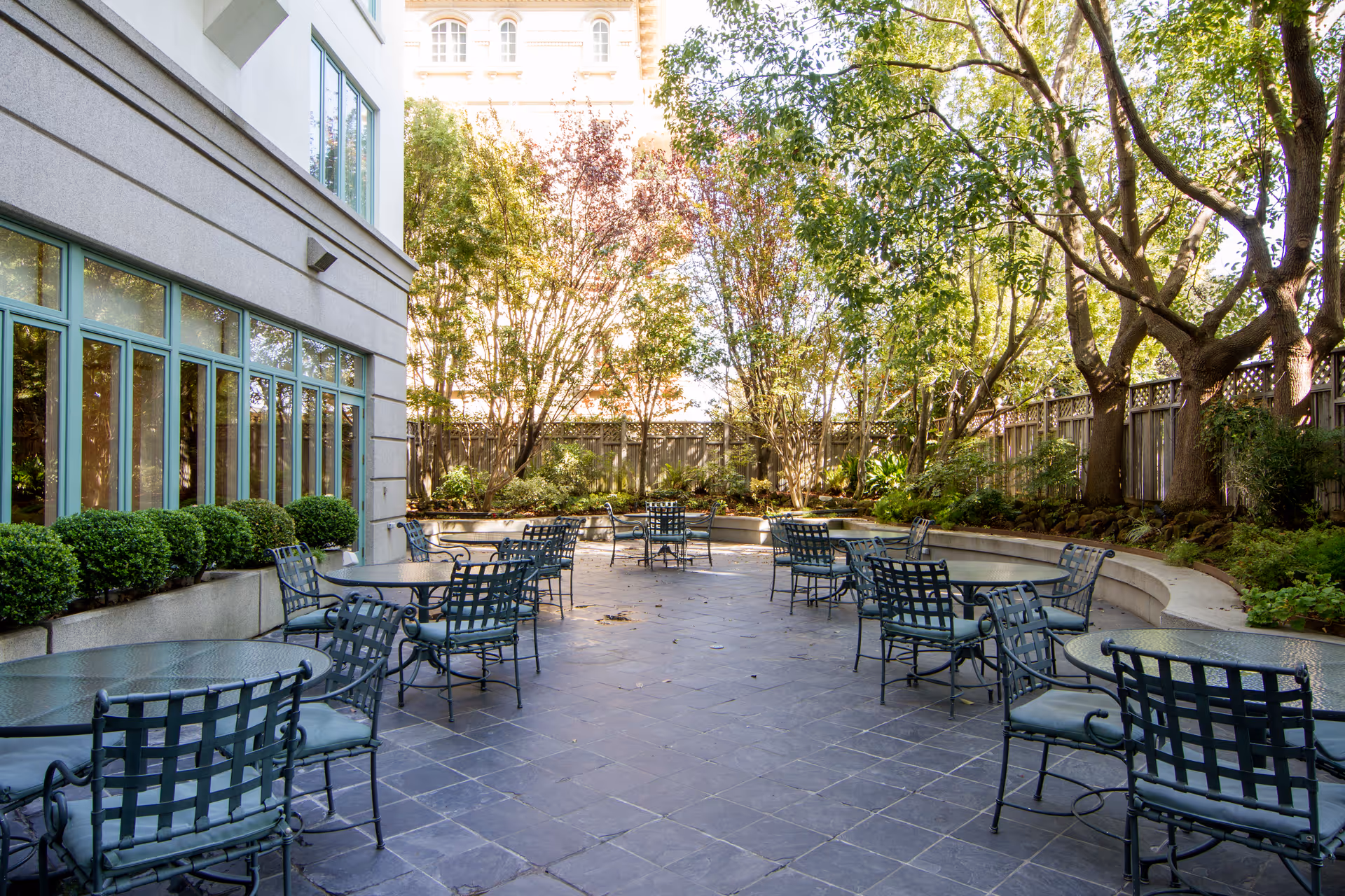Outdoor patio area at The Stratford with multiple round glass tables and metal chairs with cushions, surrounded by trees and greenery, adjacent to a building with large windows.