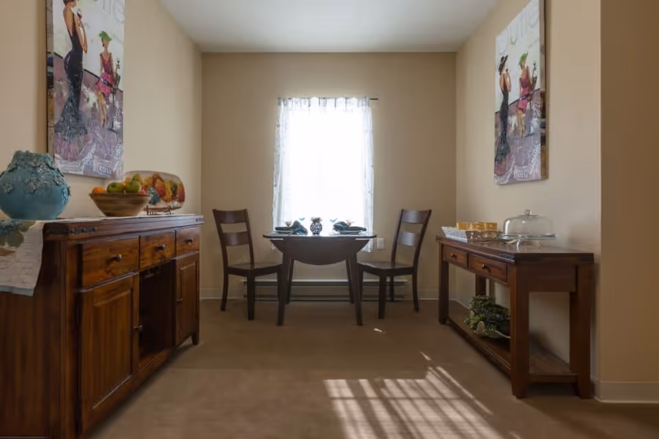A small dining area with a round wooden table set for two, positioned in front of a window with sheer white curtains. Two wooden chairs are placed on either side of the table. On each side of the room, there are wooden sideboards with decorative items including a blue vase, a bowl of fruit, and a glass cake dome. The walls are beige with two framed paintings featuring women in elegant dresses.