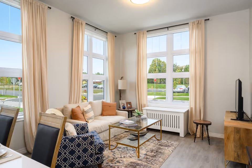 A bright living room with large windows covered by beige curtains. The room features a beige sofa with orange and patterned pillows, a blue patterned armchair, a glass coffee table with books and decor, a wooden TV stand with a flat-screen TV, and a small wooden stool. Outside the windows, a green lawn and parked cars are visible.