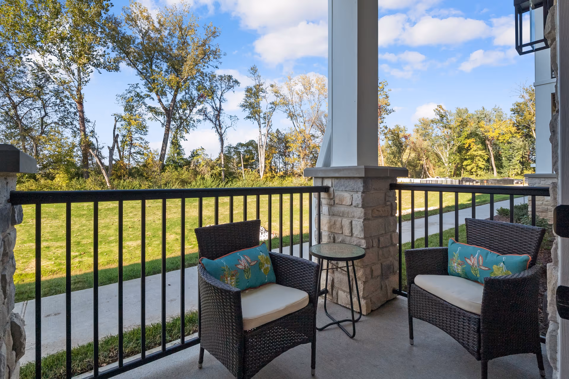 A covered outdoor patio area with two wicker chairs featuring floral cushions and a small round glass-top table between them. The patio overlooks a grassy area with trees and a clear blue sky with some clouds.