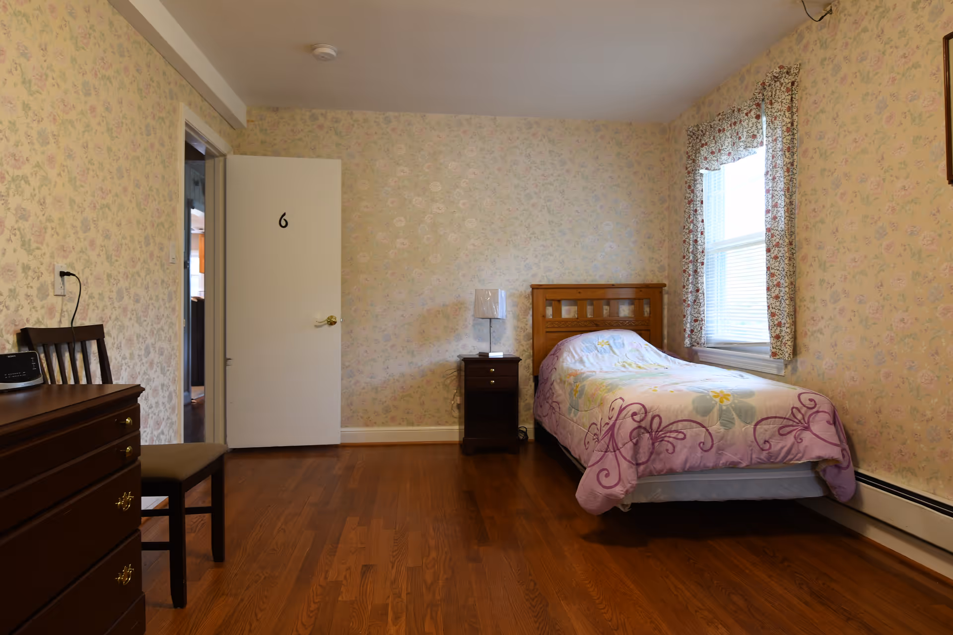 Single-occupancy bedroom with a twin bed, nightstand, dresser and floral wallpaper.