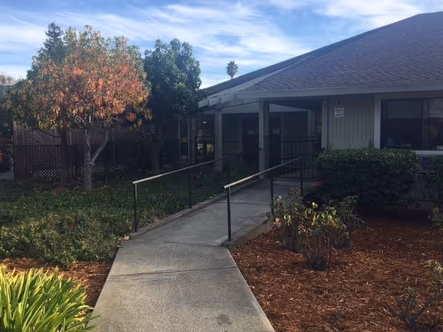 Concrete ramp with handrails leading to the entrance of a single-story building surrounded by landscaping and trees under a partly cloudy sky.