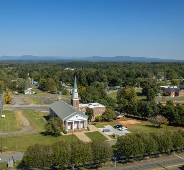 Aerial view of a church with a steeple surrounded by trees, nearby buildings, parking areas, and distant hills under a clear sky.