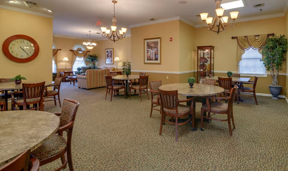 A spacious senior living common area with multiple round tables and chairs arranged for seating. The room features beige walls, carpeted floors, and several decorative plants. There are framed pictures on the walls, large windows with curtains, and ceiling light fixtures. In the background, there is a seating area with sofas and lamps.