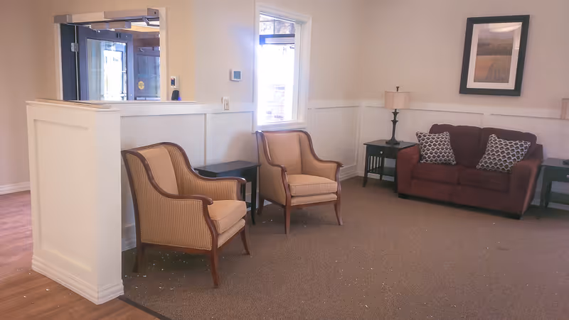 A cozy seating area in a senior living facility featuring two beige armchairs with wooden frames, a small black side table between them, a maroon loveseat with two patterned cushions, two black side tables with lamps, a framed picture on the wall, and a window letting in natural light.