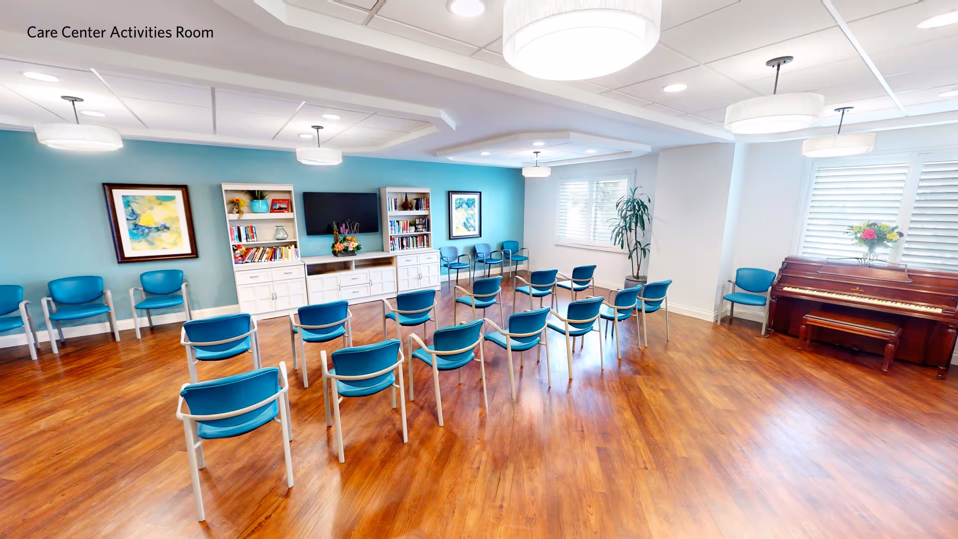 A spacious care center activities room with wooden flooring and light blue walls. The room is arranged with multiple blue chairs set in rows facing a white entertainment center with bookshelves, a TV, and decorative items. There are framed artworks on the walls, large windows with white blinds, a potted plant, and a piano with a flower vase on top.