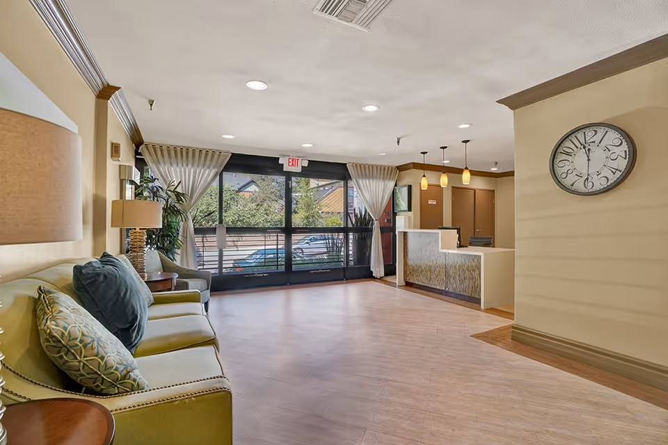 A bright and spacious senior living facility lobby with a green sofa adorned with patterned and solid pillows, a side table with a lamp, and a large clock on the wall showing 12:00. The lobby features large windows with sheer curtains, a reception desk with pendant lights above, and a view of parked cars and greenery outside.
