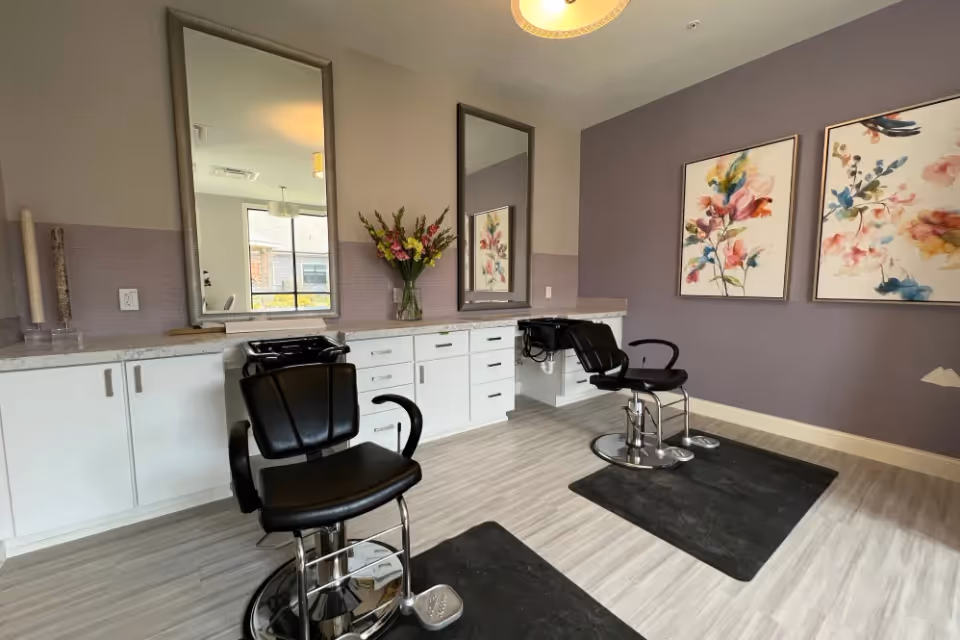 Interior salon area with two black styling chairs, large mirrors, white cabinets, and floral artwork on purple walls.