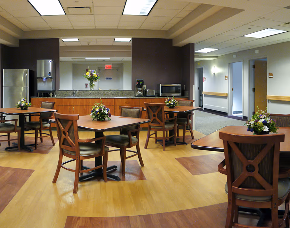 A dining area in a senior living facility with several wooden tables and chairs, each table decorated with a small floral arrangement. In the background, there is a kitchenette with a refrigerator, water dispenser, coffee maker, microwave, and a large mirror above the counter. The room has a combination of wood and carpet flooring and is well-lit with ceiling lights.