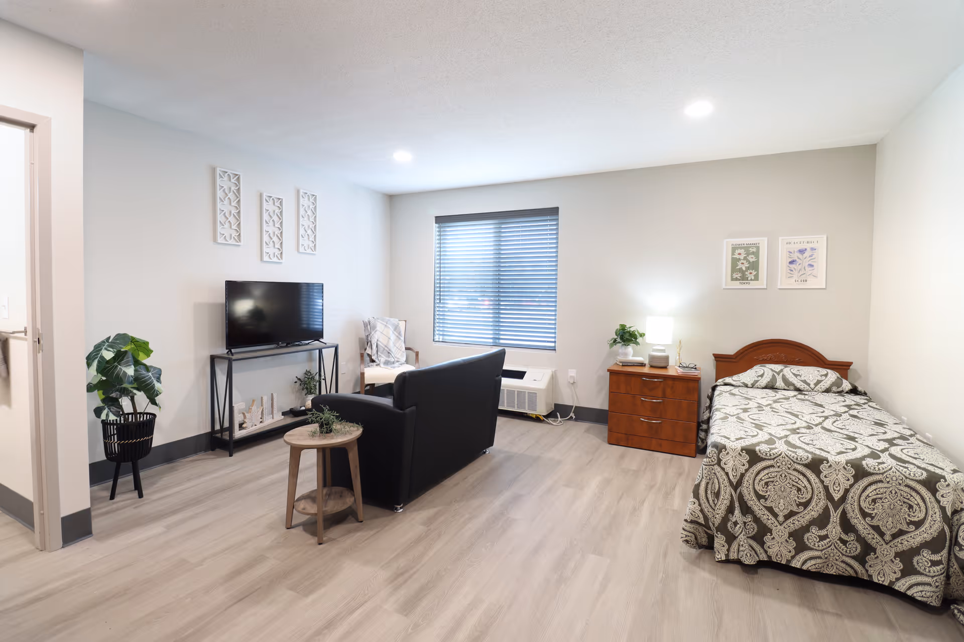 A bright and clean senior living room and bedroom area featuring a single bed with patterned bedding, a wooden nightstand with a lamp and plant, a black couch facing a TV on a stand, a small round wooden side table, a chair with a blanket, and a window with blinds. The walls are decorated with framed artwork and decorative panels, and the floor is light wood.