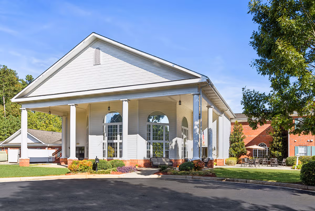 Exterior view of a senior living facility building with white siding, large white columns, and tall arched windows. There is a paved driveway in front, green grass, shrubs, and trees surrounding the building under a clear blue sky.