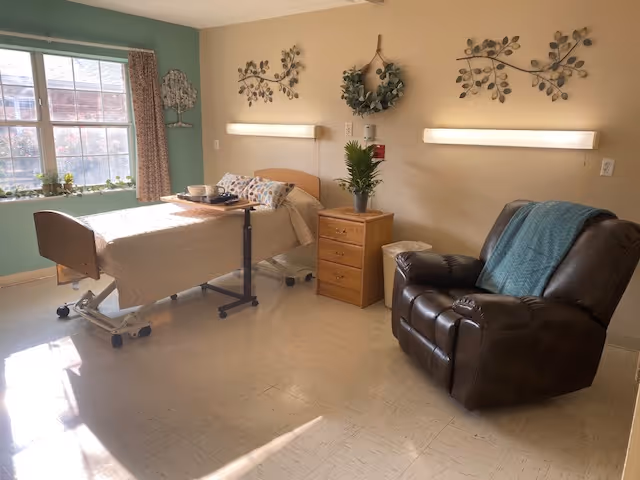 A bright, clean senior living bedroom with a hospital-style bed covered in beige bedding, a wooden nightstand with a plant, a comfortable brown recliner with a blue throw blanket, and decorative wall art including a wreath and metal leaf designs. A large window with curtains lets in natural light.