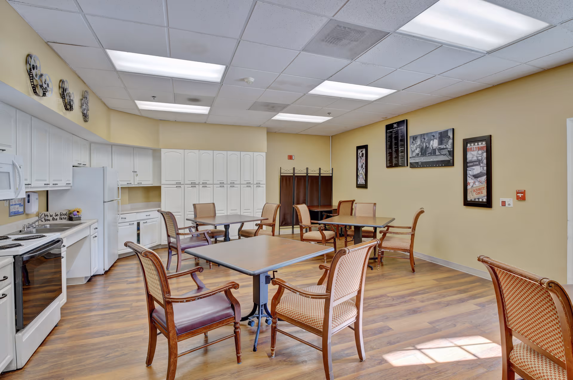 A bright and clean communal dining area with several wooden tables and chairs arranged on a wood floor. The room features a kitchen area with white cabinets, a refrigerator, stove, microwave, and dishwasher along one wall. The walls are painted light yellow and decorated with framed pictures. The ceiling has recessed fluorescent lighting.