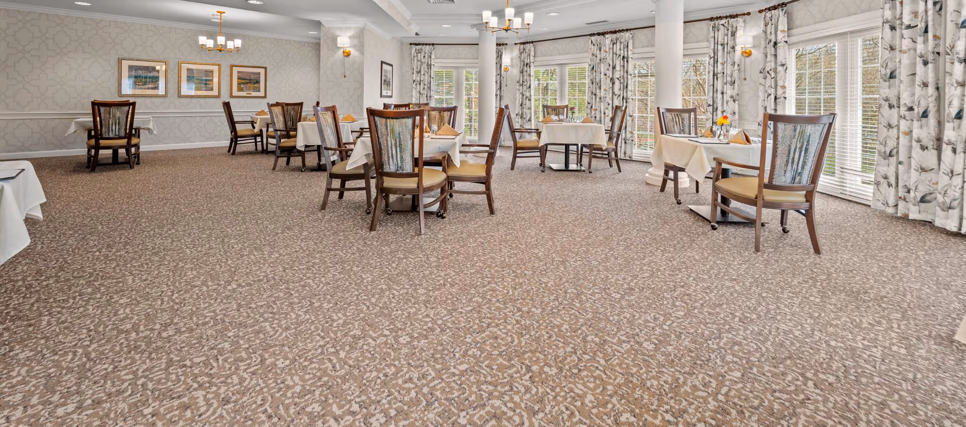 Bright dining room with multiple tables covered in white cloths, wooden chairs, chandeliers, patterned carpet, and large windows with curtains.