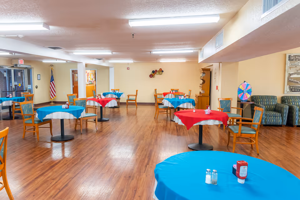 A spacious dining room with several round tables covered with red and blue tablecloths. Each table has wooden chairs with green cushions. The room has wooden flooring, beige walls, and fluorescent ceiling lights. There is an American flag near the entrance door, a colorful decorative wall piece, a small wooden cabinet, and a colorful spinning wheel on a side table. Two green patterned armchairs are placed against the right wall.