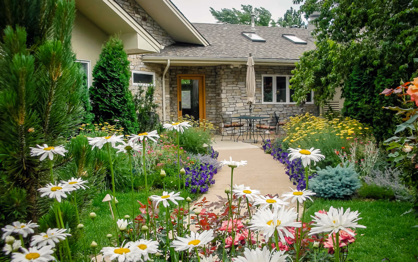 A garden pathway leading to a stone building entrance with a glass door and windows. The pathway is bordered by lush green plants, colorful flowers including white daisies, and small trees. There is an outdoor table with chairs and a closed umbrella near the entrance.