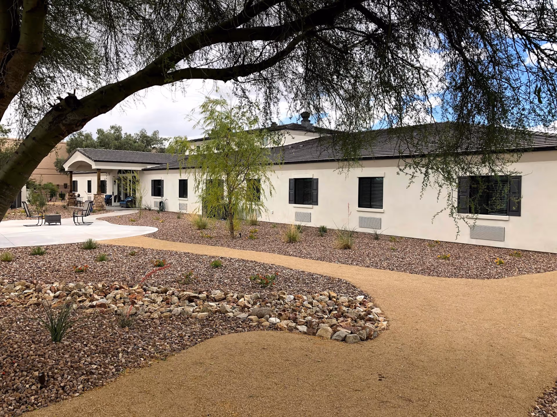 Outdoor view of a single-story assisted living facility building with beige walls and dark window shutters. The foreground features a landscaped garden with gravel, small plants, a dry creek bed made of rocks, and a winding dirt path. There are a few chairs and a fire pit on a concrete patio near the building entrance. Tree branches frame the top of the image.