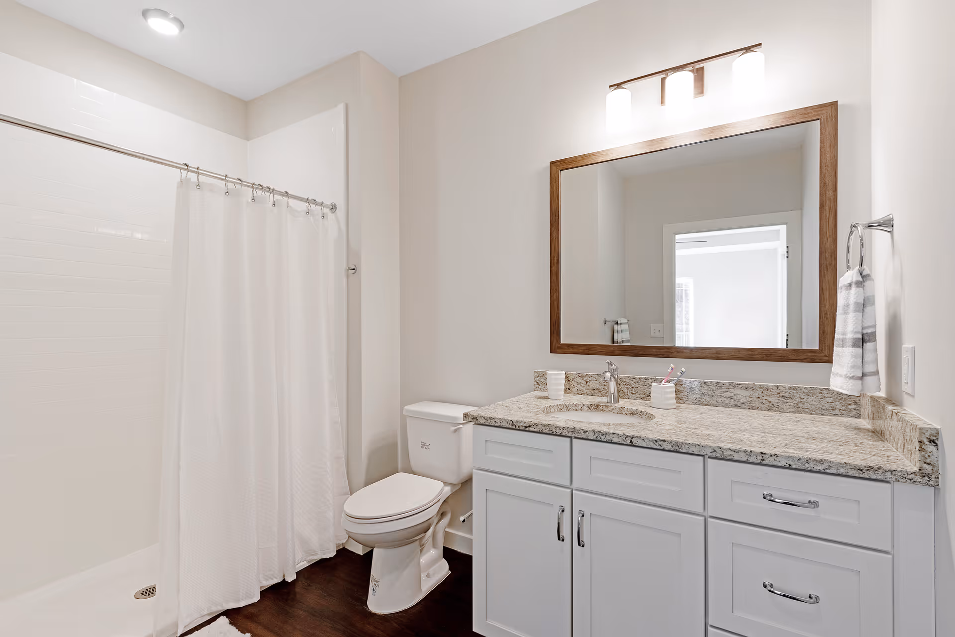 A clean and modern bathroom featuring a white shower with a curtain, a white toilet, and a granite countertop with a sink. Above the sink is a large rectangular mirror with a wooden frame and a light fixture with three bulbs. A striped hand towel hangs on a ring next to the sink.