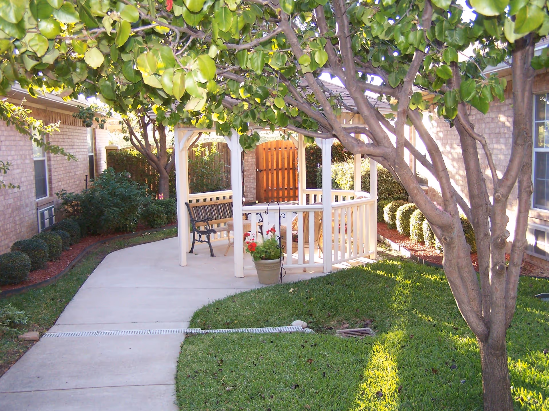 A peaceful outdoor garden area at Sodalis Flower Mound featuring a white gazebo with a bench and chairs inside, surrounded by green grass, trees, and neatly trimmed bushes along a concrete pathway.