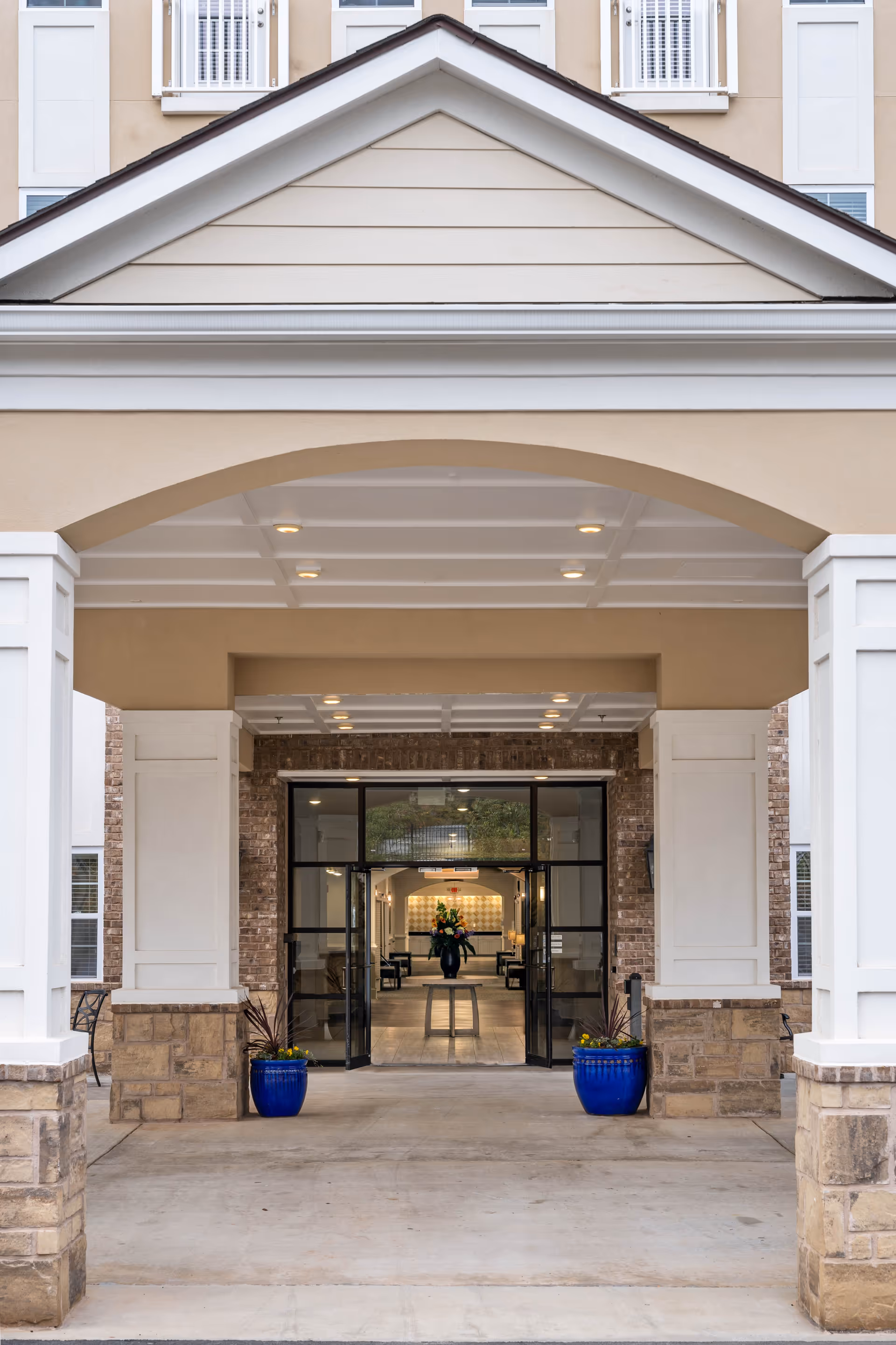 Entrance of a senior living facility with a covered driveway supported by white columns and stone bases. Two blue planters with plants flank the entrance, leading to glass double doors that open into a well-lit interior with a table and flower arrangement visible inside.