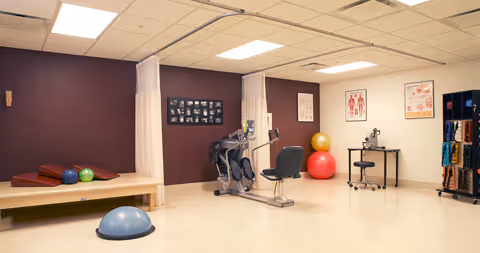 A physical therapy or rehabilitation room with exercise equipment including a stationary bike, therapy balls, balance trainers, and a therapy table with cushions. The walls have anatomical charts and a photo collage, and there is a rolling cart with various therapy tools.