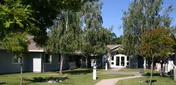Exterior view of a single-story retirement living community building surrounded by green grass and several trees under a clear blue sky.