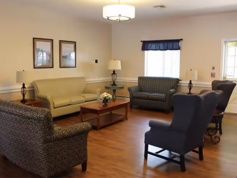 Communal living room with sofas, armchairs, a coffee table, lamps, and wood flooring.