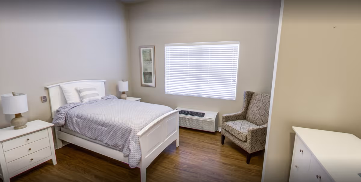A simple, clean bedroom with a white wooden bed frame, a bed with gray patterned bedding and pillows, two white nightstands each with a lamp, a patterned armchair near a window with closed blinds, a framed artwork on the wall, and a white dresser on the right side. The floor is wooden and the walls are painted beige.
