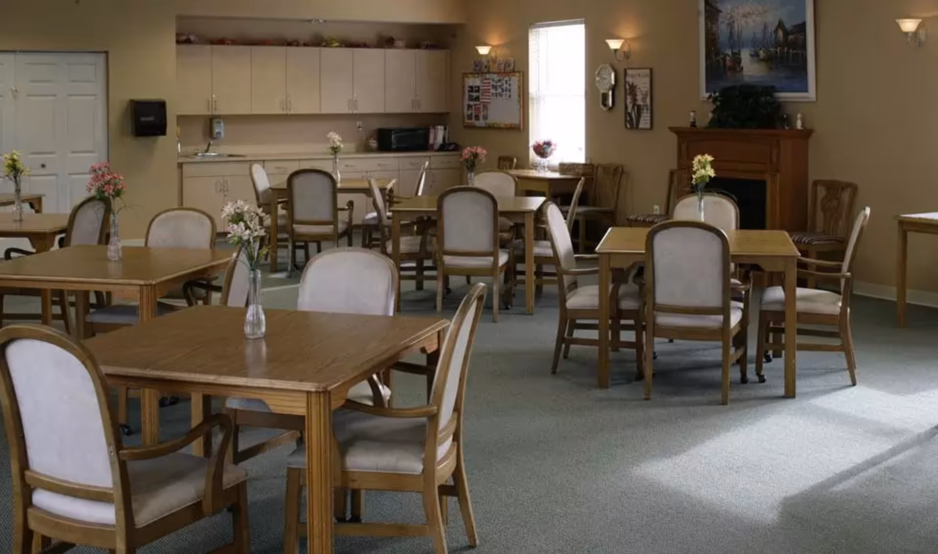 A dining room with multiple wooden tables and cushioned chairs arranged neatly. Each table has a small vase with flowers. The room has beige walls, a carpeted floor, a fireplace with a painting above it, and a window letting in natural light. There are cabinets and a countertop with a microwave in the background.
