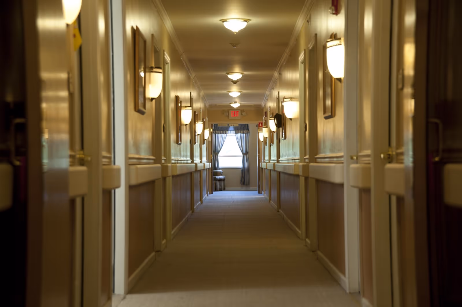 A long, well-lit hallway in an assisted living facility with multiple doors on both sides, wall-mounted lights, and a window with curtains at the far end.