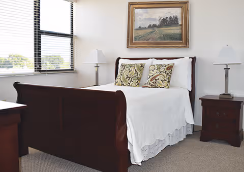 A bedroom with a wooden bed frame, white bedding, and two decorative pillows. There are two matching wooden nightstands on either side of the bed, each with a lamp. A framed landscape painting hangs on the wall above the bed. A window with blinds is on the left side of the room.