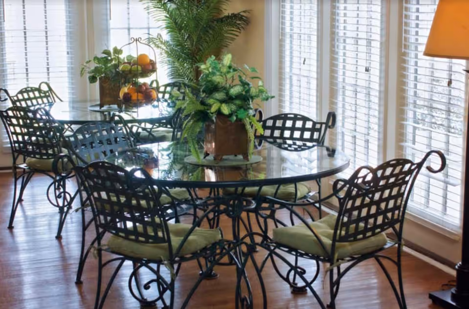 A dining area with a glass-top table surrounded by six wrought iron chairs with green cushions. The table has a centerpiece with green plants and a tiered fruit basket. The room has large windows with white blinds allowing natural light to fill the space, and a floor lamp is visible on the right side.