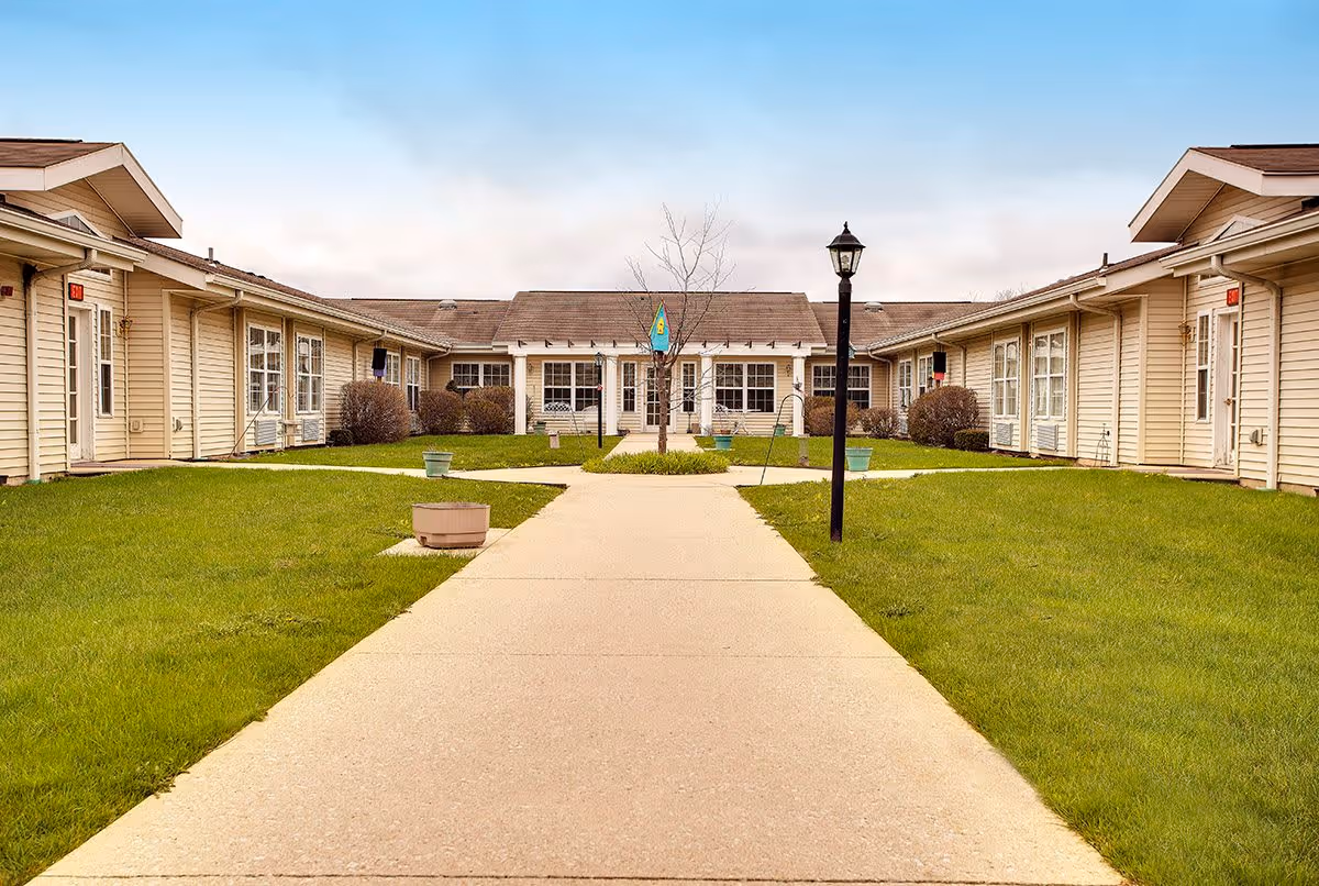 A courtyard area at Trustwell Living at Settlers Place featuring a concrete walkway leading to a central building entrance. The courtyard is surrounded by single-story beige buildings with multiple windows and doors. There is a lamppost along the walkway, some small potted plants, and a flag hanging near the entrance. The sky is clear with a few clouds.