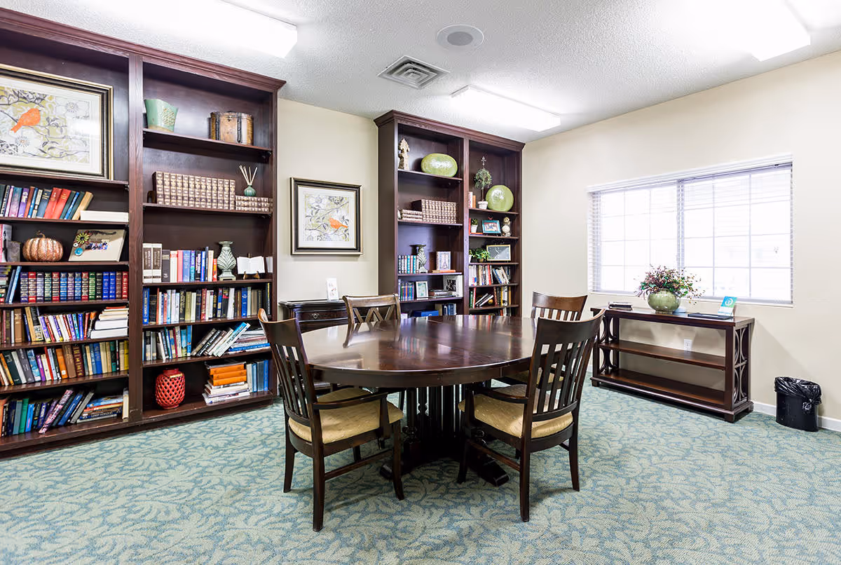 A bright room with a round wooden table surrounded by four chairs with cushioned seats. Behind the table are two large dark wood bookshelves filled with books, decorative items, and framed artwork. A window with blinds lets in natural light, and a small console table with a flower arrangement is positioned beneath the window. The room has a patterned carpet and white walls.